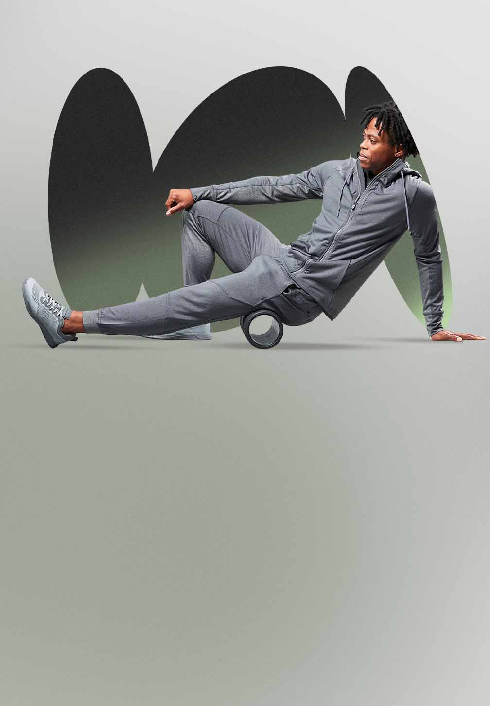 An African American man wearing a Hylete Flexion II Jacket and Flexion II Jogger in a gray color is using a black foam roller on the back of his upper leg. He is on a light gray/green floor with a darker gray/green abstract background.