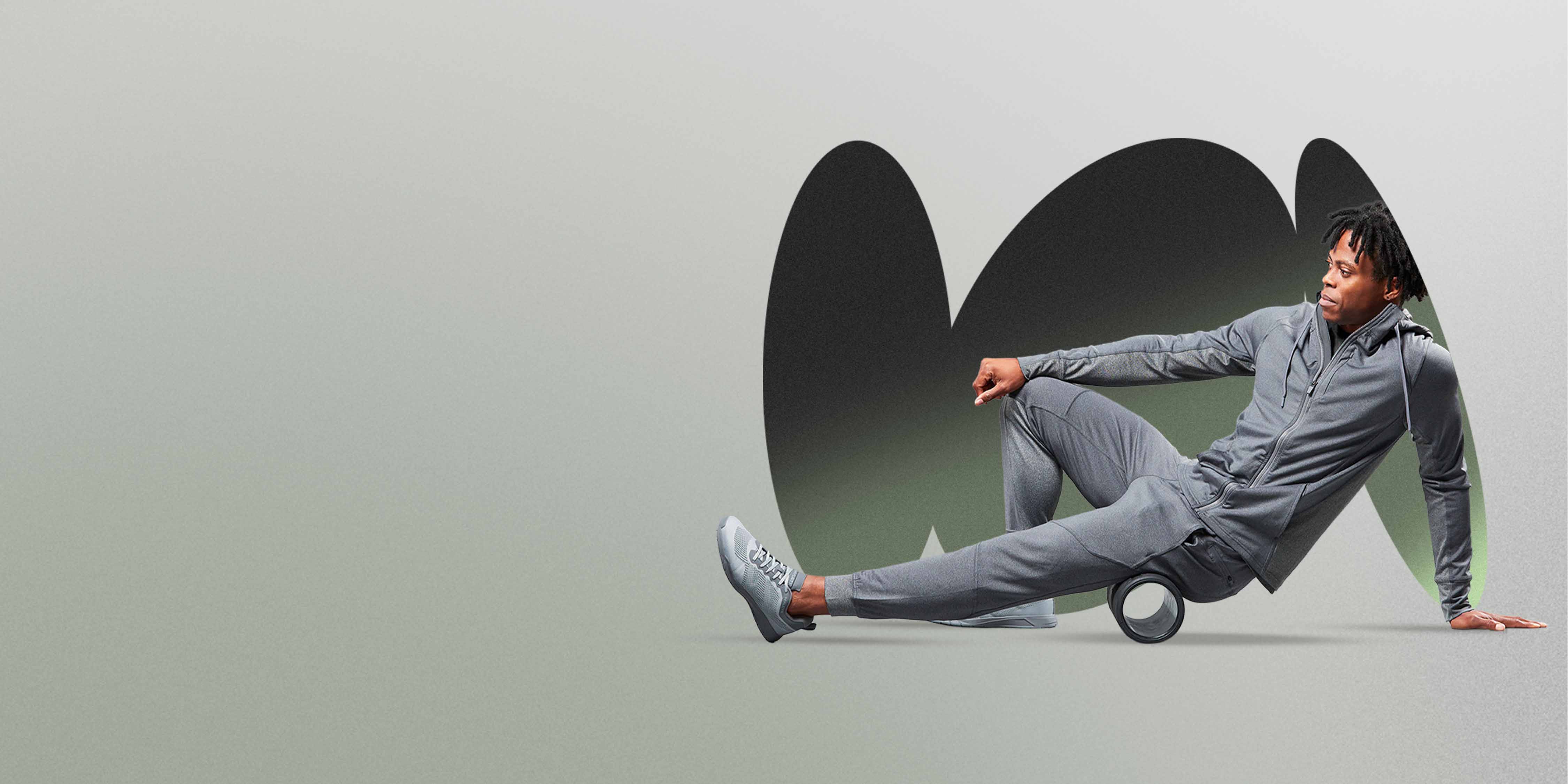 An African American man wearing a Hylete Flexion II Jacket and Flexion II Jogger in a gray color is using a black foam roller on the back of his upper leg. He is on a light gray/green floor with a darker gray/green abstract background.