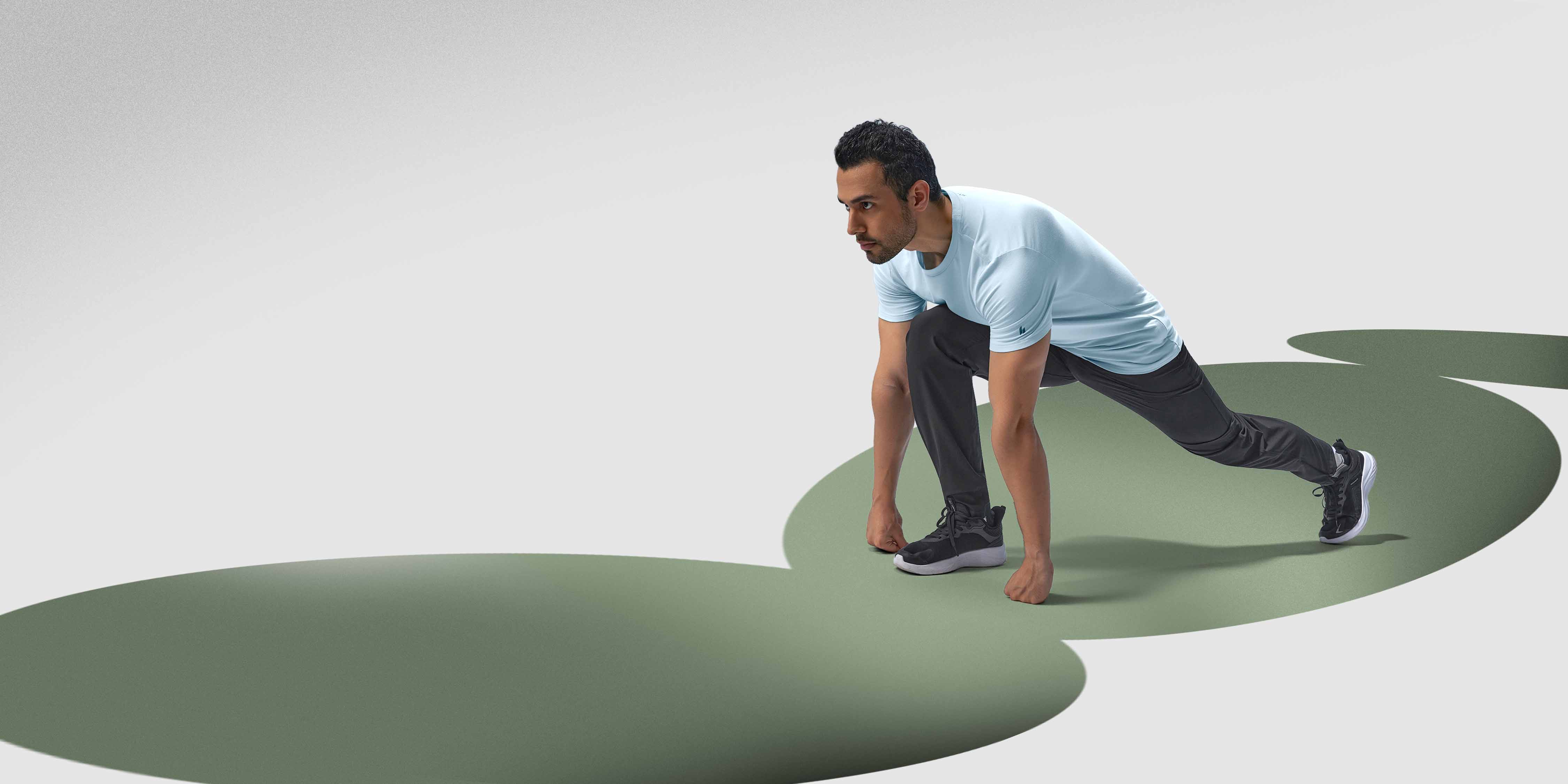 A man wearing black Hylete Cadence run pant and a light blue HY-Blend Crew Tee performs a forward lunge. His right leg is extended back, and his left leg is bent at a 90-degree angle, with his foot flat on the ground. He is focused and looking straight ahead.