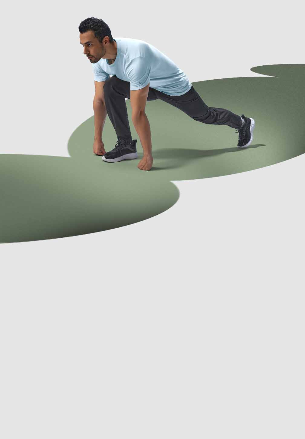 A man wearing black Hylete Cadence run pant and a light blue HY-Blend Crew Tee performs a forward lunge. His right leg is extended back, and his left leg is bent at a 90-degree angle, with his foot flat on the ground. He is focused and looking straight ahead.