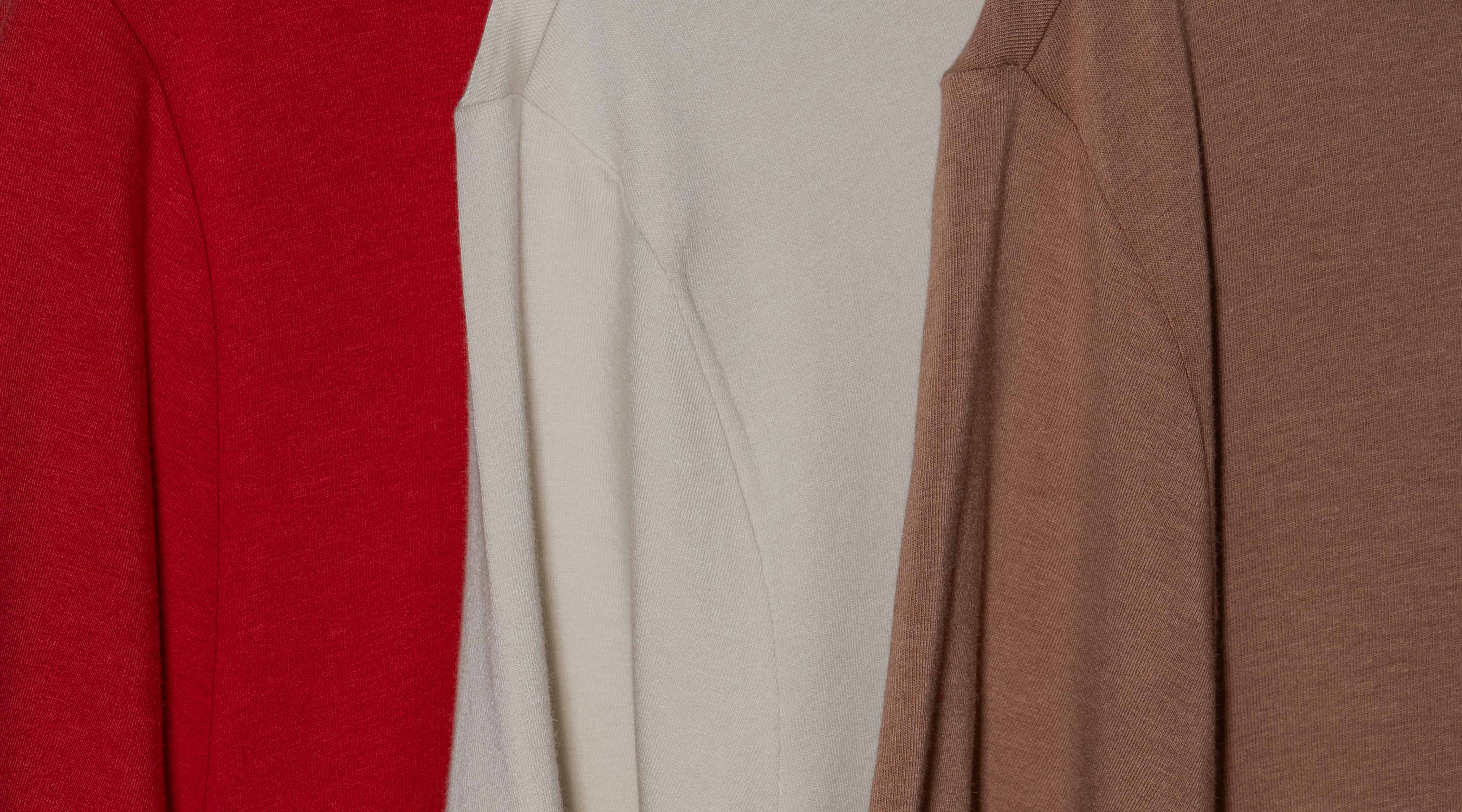 Close-up shot shows three Hylete HY-Blend crew tees hanging side by side. From left to right, the colors are a crimson red, a bone white, and a raw umber.