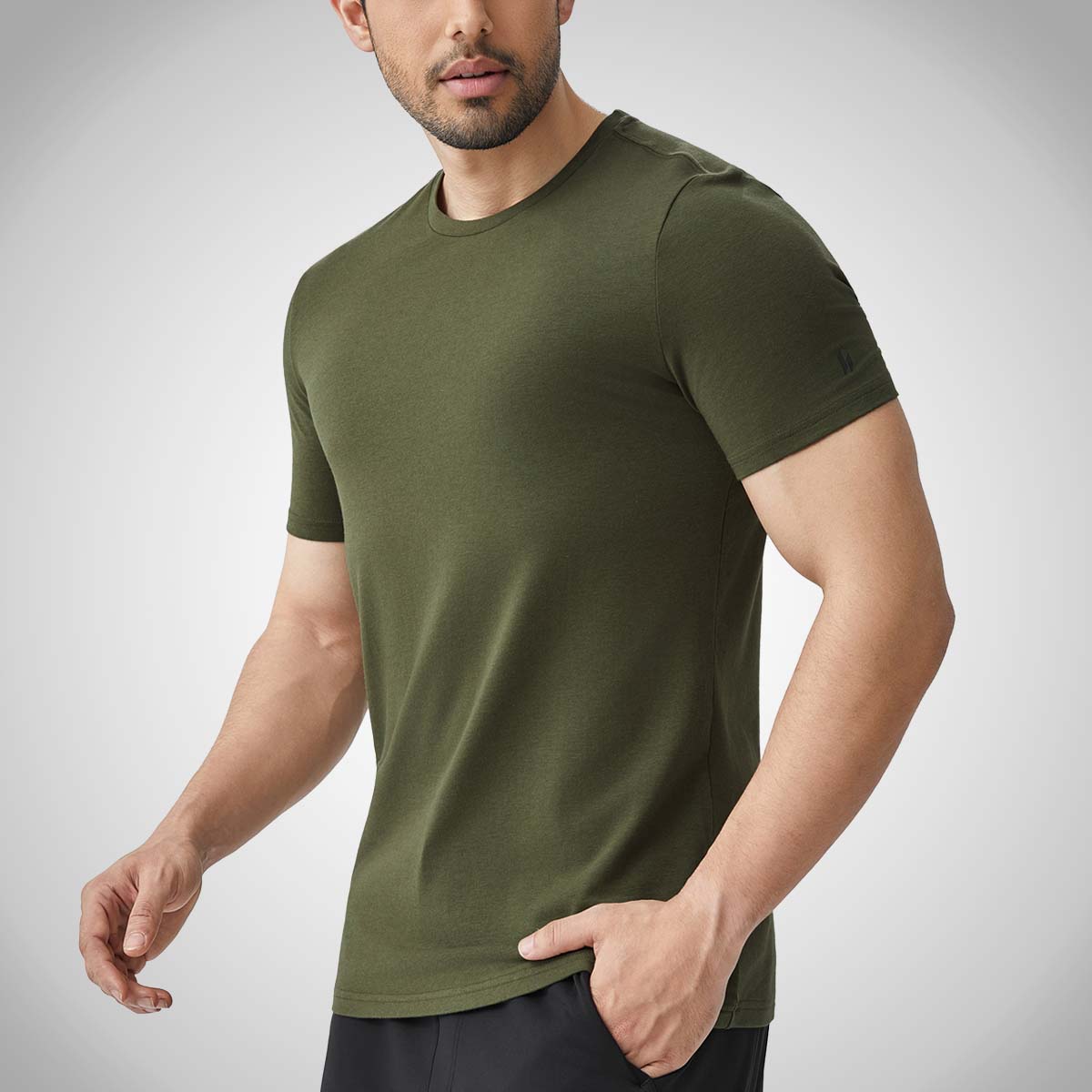 A man, shown from the chest down and facing left, wearing an olive green HYLETE HY-Blend Crew Tee, and black shorts. His hand is resting in his pocket.