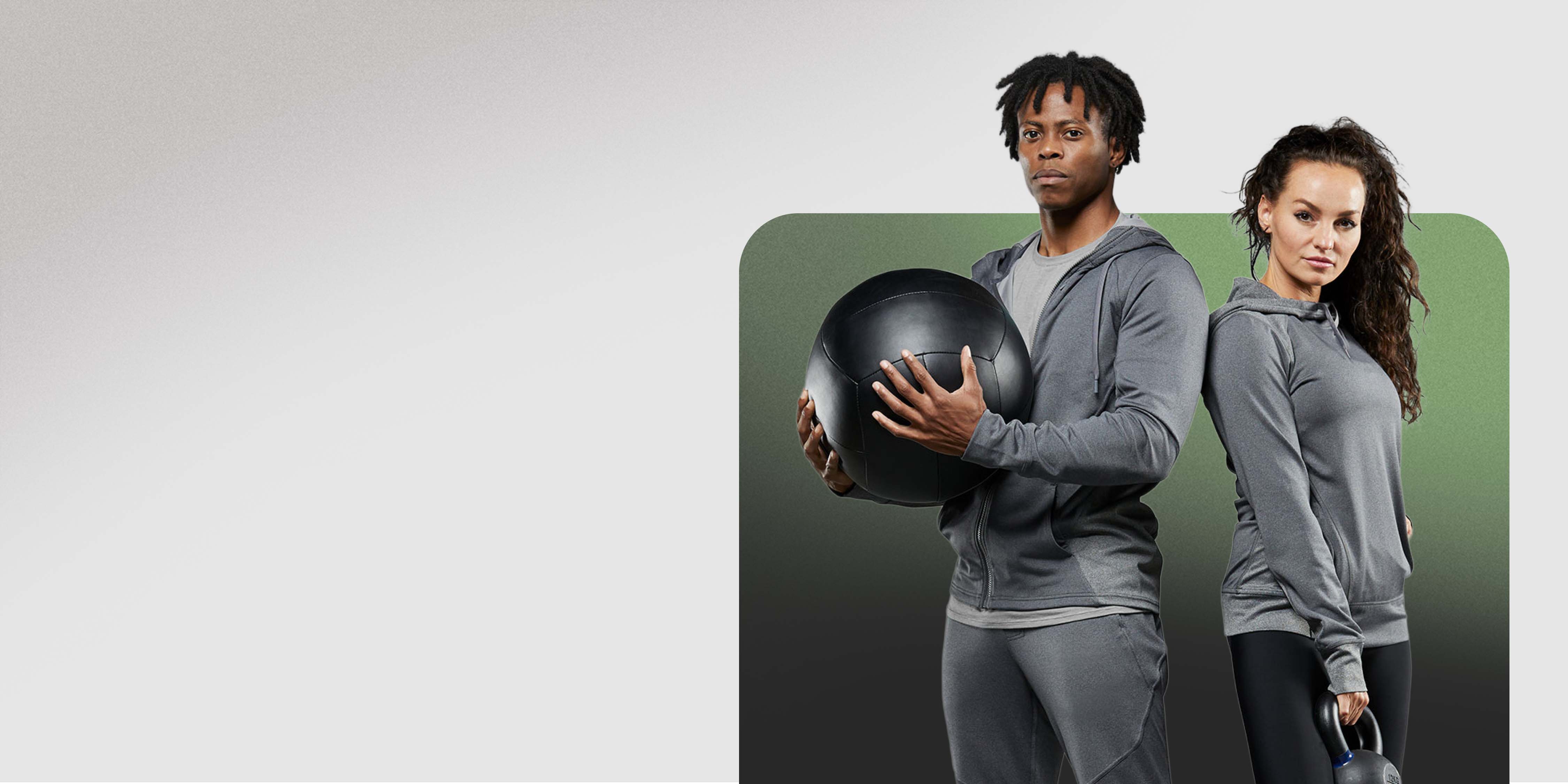 A man and a woman in Hylete gear stand in a gym. The man on the left is holding a black medicine ball, and the woman on the right is holding a kettlebell.