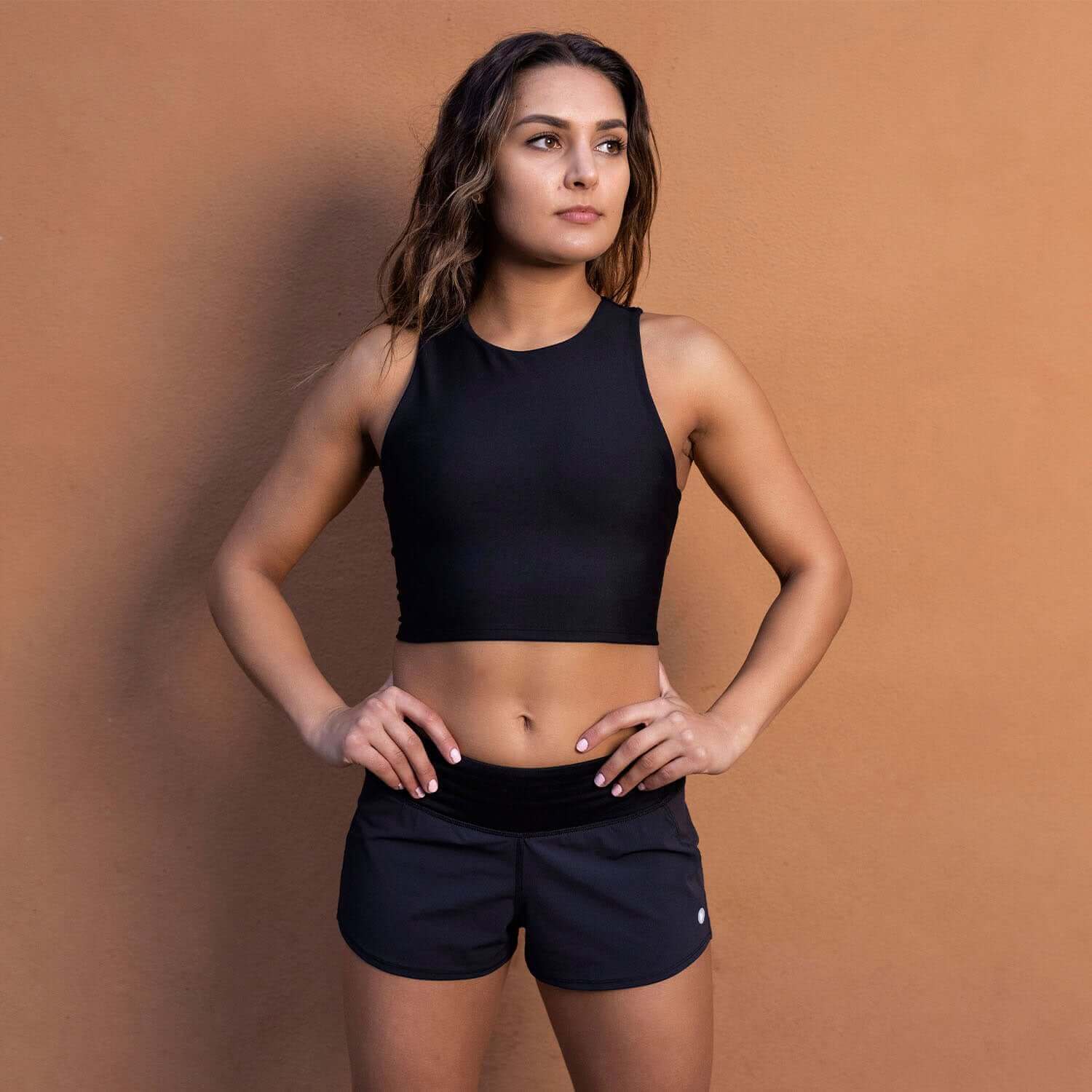 Woman wearing Vita Short Black athletic shorts and crop top for running or HIIT workout against a brown background.