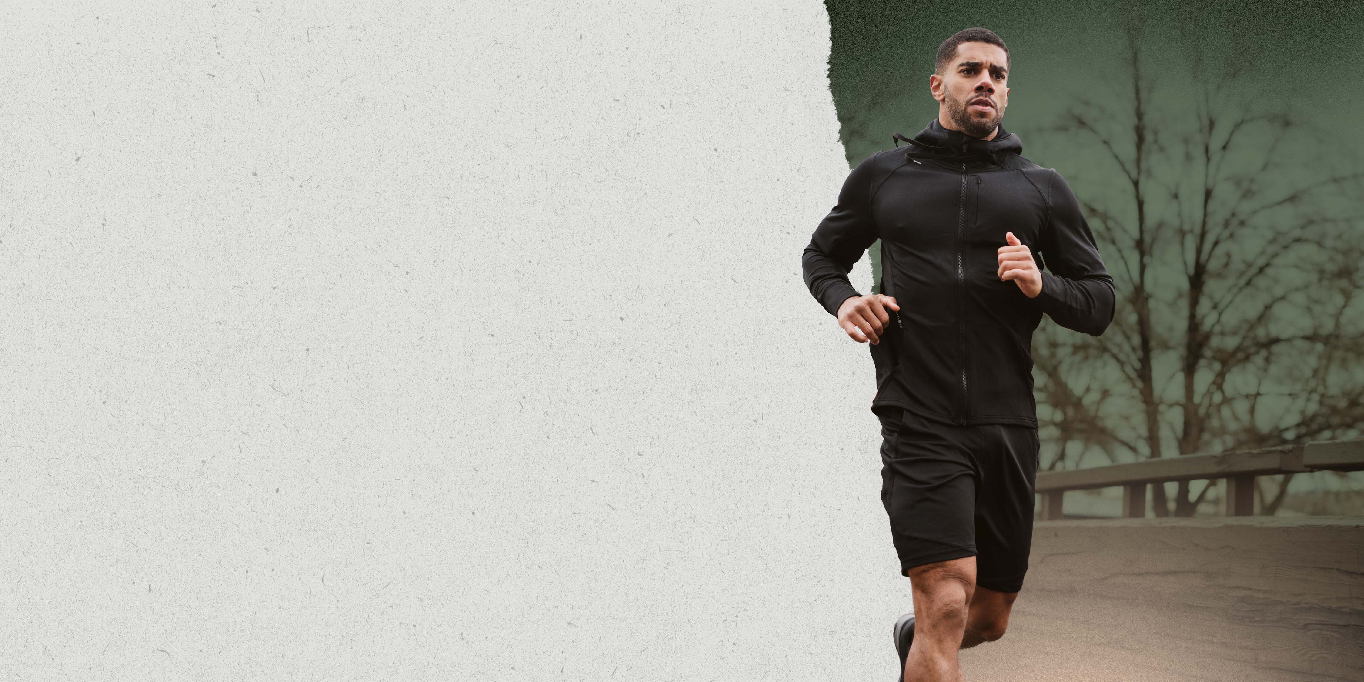 A determined male runner wearing the Hylete Flexion II Jacket and Flexion II Short in black color runs outdoors in cold weather, with bare trees and a snowy foreground.