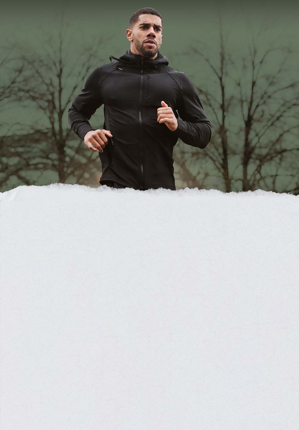 A determined male runner wearing the Hylete Flexion II Jacket in black color runs outdoors in cold weather, with bare trees and a snowy foreground.